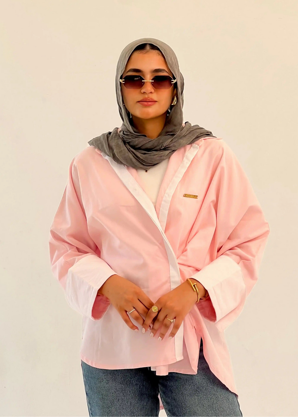 Person wearing a pink shirt, gray hijab, and sunglasses against a beige background